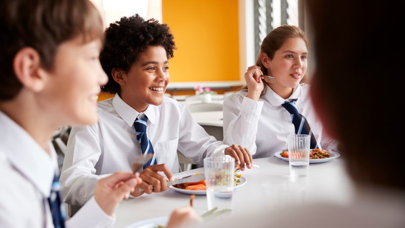 Group Of High School Students Wearing Uniform Sitting Around Table And Eating Lunch In Cafeteria